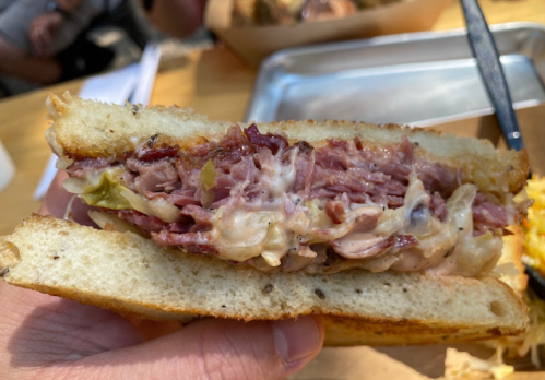 A close-up of a hand holding a sandwich filled with layers of meat, cheese, and sauerkraut between two slices of toasted bread.