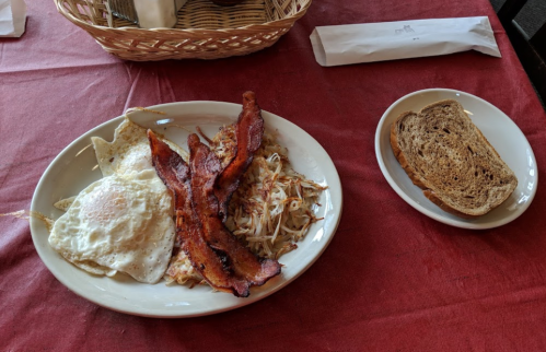 A plate of two fried eggs, crispy bacon, coleslaw, and a side of whole grain toast on a red tablecloth.