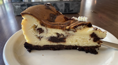 A slice of cheesecake with a chocolate swirl on a white plate, resting on a wooden table.
