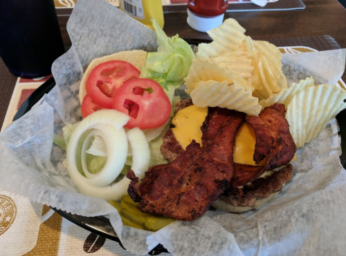 A burger with bacon and cheese, served with lettuce, tomato, onion, pickles, and potato chips in a basket.