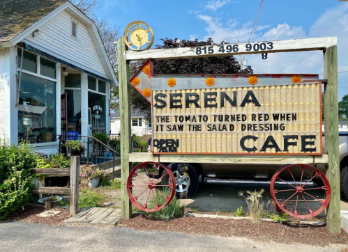 A rustic café sign reads "Serena: The tomato turned red when it saw the salad dressing. Open." with a charming exterior in the background.