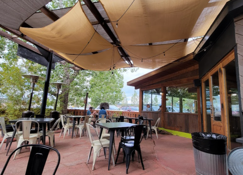 Outdoor dining area with tables and chairs, shaded by a canopy, surrounded by trees and a casual atmosphere.