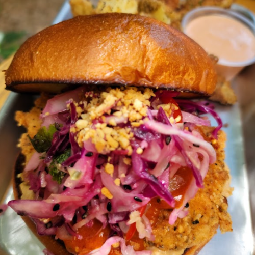 A close-up of a crispy chicken sandwich topped with purple slaw, tomatoes, and a side of dipping sauce.