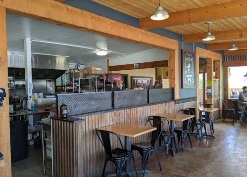 Interior of a casual restaurant featuring wooden beams, metal chairs, and an open kitchen area.