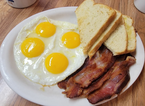 A plate with three sunny-side-up eggs, crispy bacon strips, and slices of toasted bread on a wooden table.