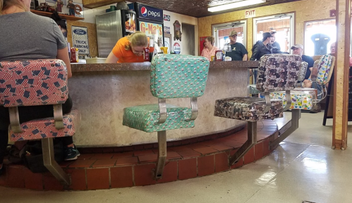 A retro diner with colorful patterned stools around a circular counter, and patrons in the background.