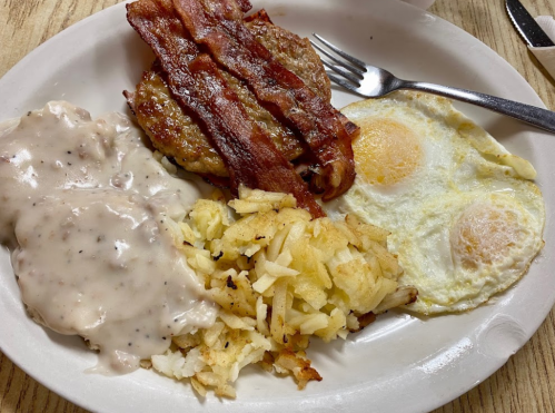 A plate of breakfast featuring bacon, sausage patty, fried eggs, hash browns, and creamy gravy.