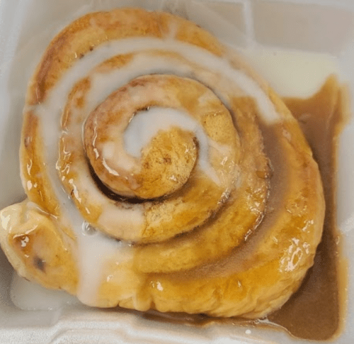 A freshly baked cinnamon roll drizzled with icing, resting in a white takeout container.