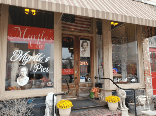 A quaint pie shop with a welcoming entrance, featuring a sign that reads "Myrtle's Pie" and colorful decorations.