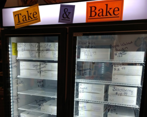 A refrigerated display case labeled "Take & Bake" with various quiches and desserts, each with prices written on boxes.
