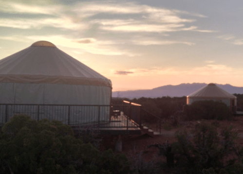 Two yurts sit on a wooden deck at sunset, with mountains in the background and a colorful sky.