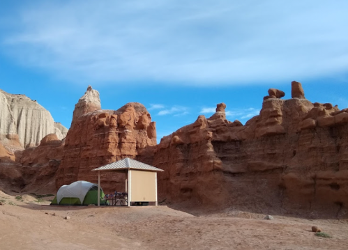 A green tent beside a small shelter, surrounded by towering red rock formations under a blue sky.