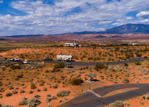 Aerial view of a desert landscape with RVs parked, surrounded by mountains and scattered vegetation under a cloudy sky.