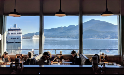 A restaurant with large windows overlooking a calm bay, mountains in the background, and diners at tables.