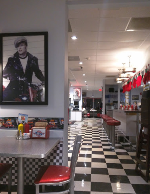 A retro diner interior with checkered floors, red stools, and a large black-and-white photo on the wall.