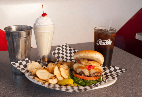 A burger with lettuce, tomato, and pineapple, served with chips, a milkshake, and a glass of cola on a diner table.