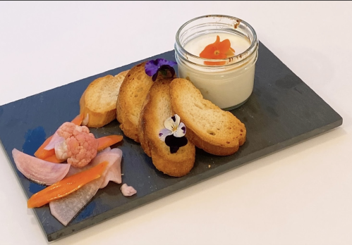 A slate platter with toasted bread, colorful vegetables, and a small jar of creamy dip, garnished with edible flowers.