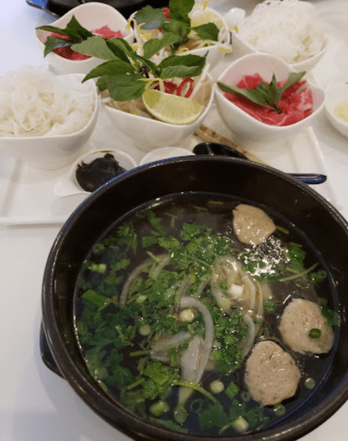 A bowl of soup with meatballs and herbs, accompanied by fresh vegetables and noodles in separate bowls.