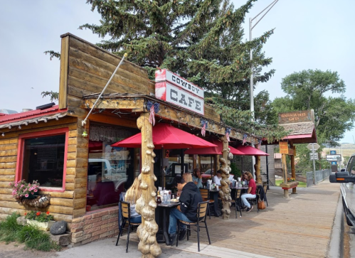 A rustic café with a wooden exterior, outdoor seating, and red umbrellas, surrounded by trees and a sidewalk.