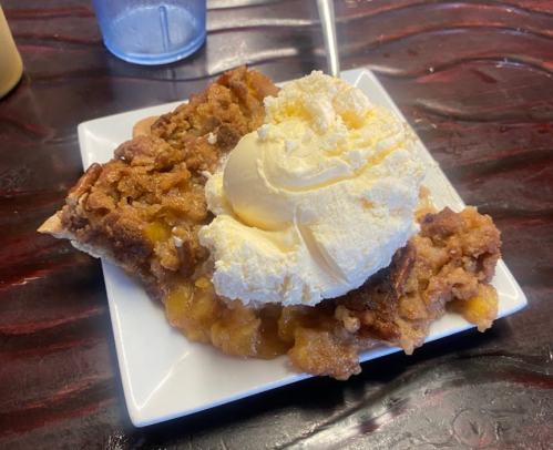 A slice of warm peach cobbler topped with a scoop of vanilla ice cream on a white plate.