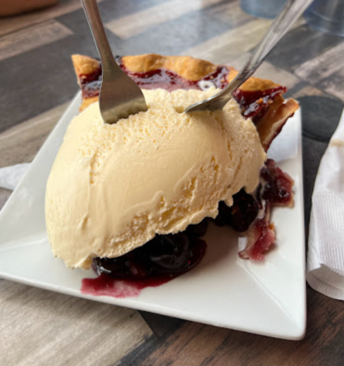 A slice of pie topped with a scoop of vanilla ice cream and drizzled with berry sauce on a white plate.