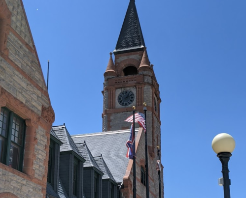 Historic clock tower with a pointed roof, flanked by flags, against a clear blue sky.