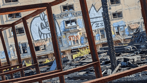 View through a rusted frame of a burned building, featuring graffiti and remnants of charred structures.