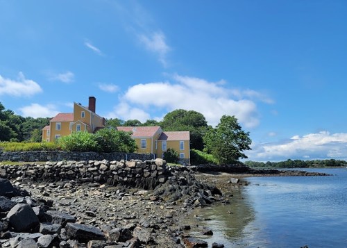 A yellow house on a rocky shore, surrounded by greenery and under a blue sky with scattered clouds.