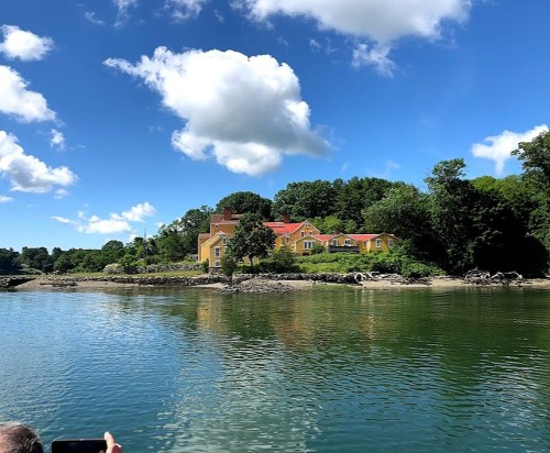 A colorful house on a hillside by a calm river, surrounded by lush greenery and blue skies with fluffy clouds.