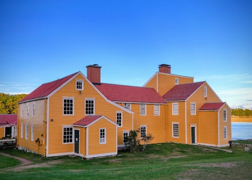 A bright yellow house with red roofs, surrounded by green grass and a clear blue sky, near a body of water.