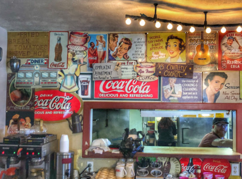 A retro diner interior with vintage Coca-Cola signs, posters, and a glimpse of the kitchen area.