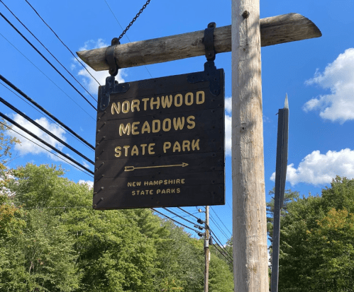 Sign for Northwood Meadows State Park in New Hampshire, hanging from a wooden post against a blue sky.