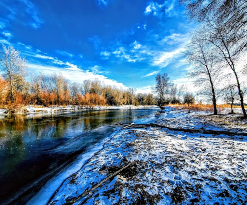 A serene winter landscape featuring a river, snow-covered banks, and trees under a bright blue sky.