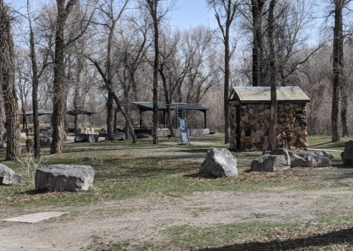A park scene featuring stone structures, large rocks, and trees, with picnic areas and a swing set in the background.