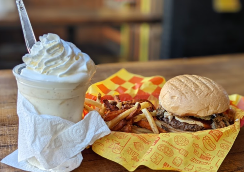 A burger with fries and a milkshake on a wooden table, served in a colorful paper basket.