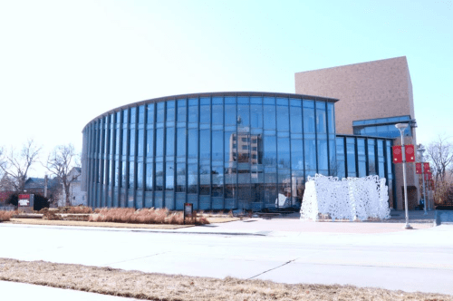 Modern glass building with a curved facade, surrounded by grass and trees, on a clear day.
