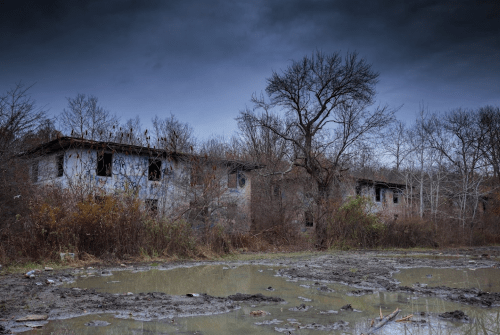 Abandoned buildings surrounded by overgrown vegetation and a muddy area under a dark, cloudy sky.