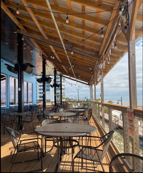 A spacious outdoor deck with tables and chairs, string lights, and a view of the water and buildings in the background.