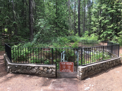 A fenced area in a forest with a sign indicating tour hours and a stone wall surrounding a natural feature.