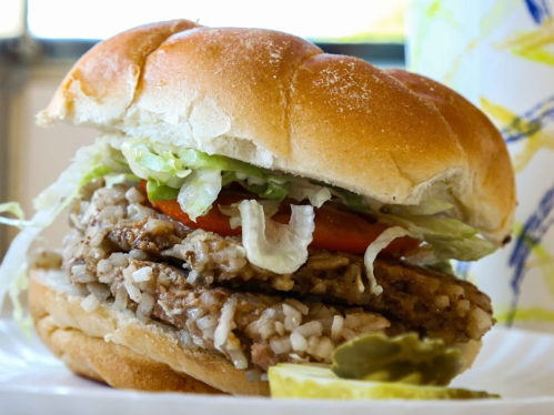 A close-up of a sandwich with a meat patty, lettuce, tomato, and rice, served on a soft bun with a slice of lime.