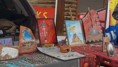 Colorful artwork and jewelry displayed on a table, featuring paintings and handcrafted items in a market setting.