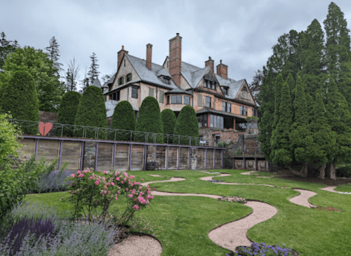 A large, elegant mansion surrounded by lush greenery and a landscaped garden with winding paths and colorful flowers.