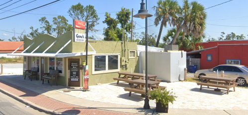 A small green restaurant with outdoor seating, surrounded by palm trees and a parking area, on a sunny day.