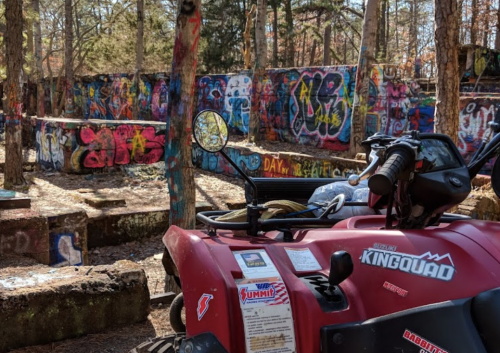 A red ATV parked in front of a colorful graffiti-covered wall in a wooded area.