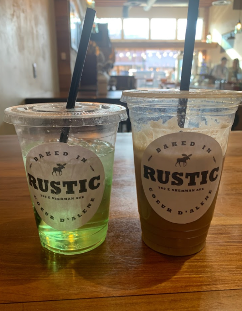 Two drinks on a wooden table, one green and one iced coffee, both in clear cups with "Rustic" labels.