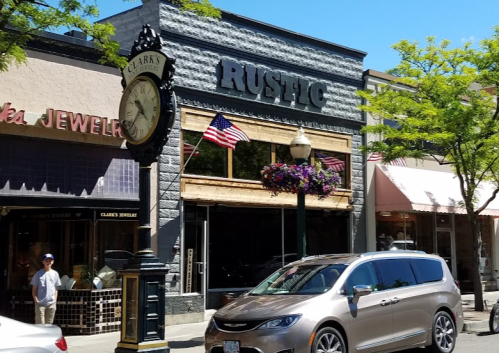 A street view of a shop named "Rustic" with American flags, a clock, and parked cars in a sunny setting.