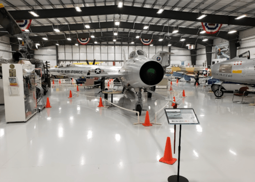 A spacious aircraft hangar featuring various planes, including a U.S. Air Force jet, with safety cones and informational signs.