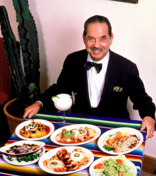A smiling man in a tuxedo presents a variety of colorful dishes on plates, with a cocktail beside him.