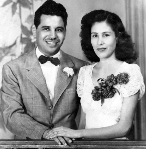 A vintage black-and-white photo of a smiling couple, dressed elegantly, with floral accents and a soft background.