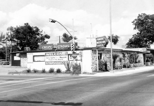 Black and white photo of a roadside restaurant with signs for crispy tostadas and steak, surrounded by greenery.